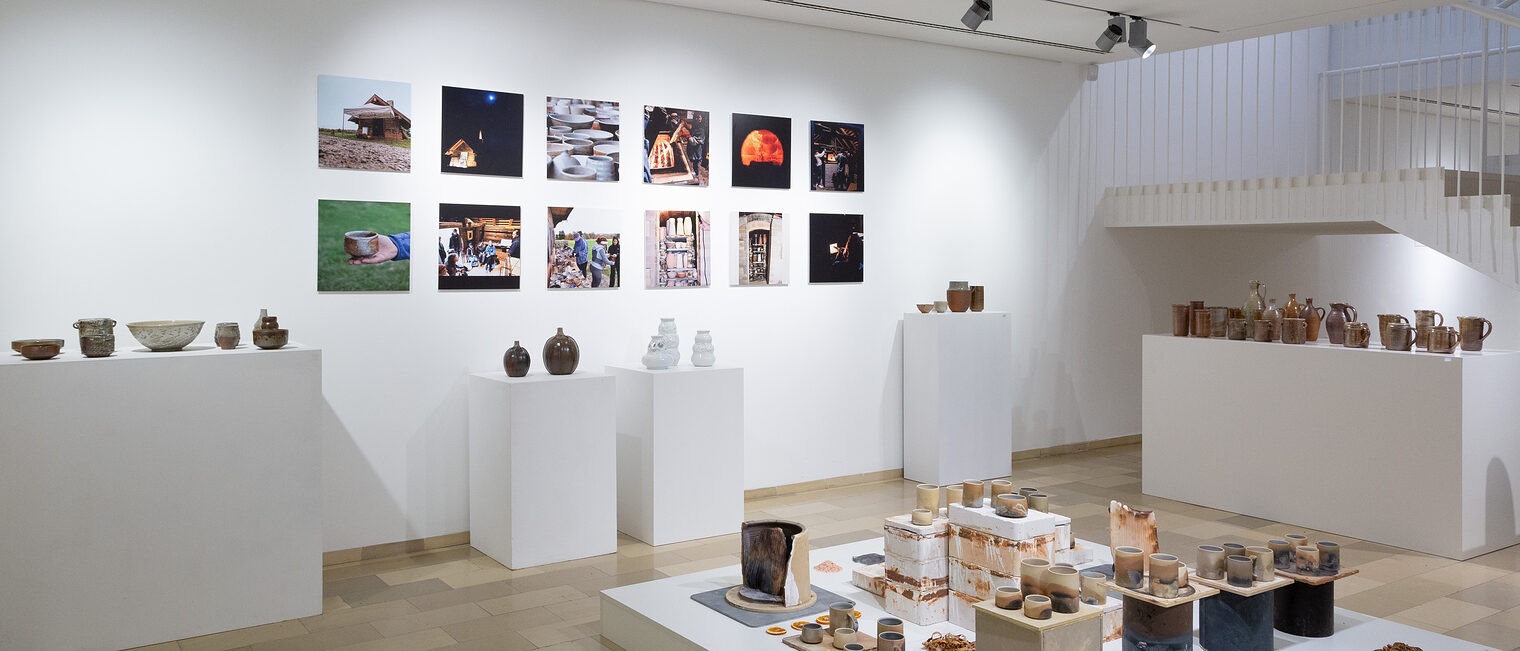 Säulen und ein Podest mit Gefäßen, Krügen, Vasen, Bechern aus Keramik. Im Hintergrund zwölf Bilder mit Herstellungsprozessen.