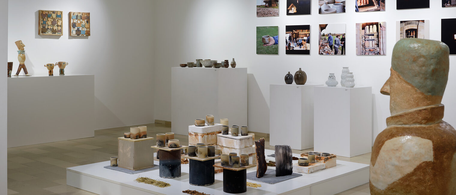 Blick in die Ausstellung. Im Vordergrund Keramikfigur, dahinter Bilder vom Herstellungsprozess und Keramikobjekte auf Säulen.