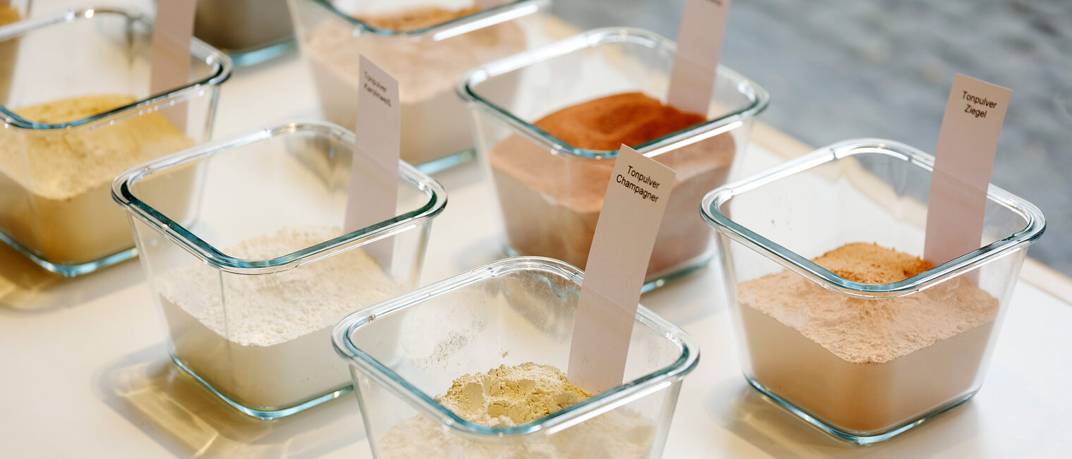 Am Schaufenster der Galerie Handwerk ein Tisch mit Farbpigmenten in gedeckter Farbe in offenen Glasbehältern.