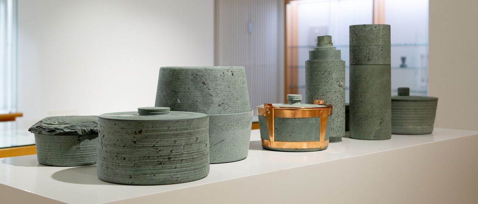 Verschiedene Steingefäße, mal in Topfform mit Deckel, mal als Vase in Steingrau auf einem Ausstellungstisch