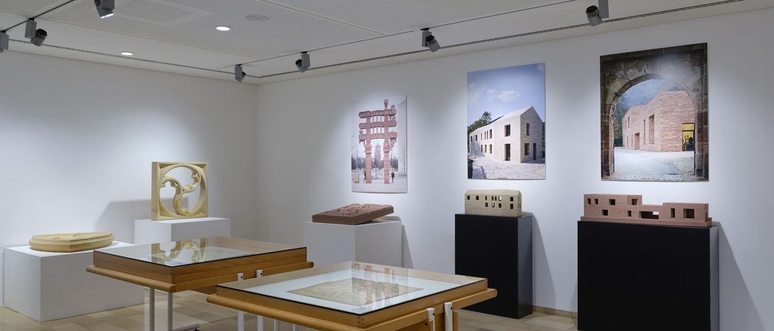 Impressionen der Steinausstellung. Im Hintergrund Fotoaufnahmen mit Steingebäuden, darunter Modelle von Häusern aus Stein.