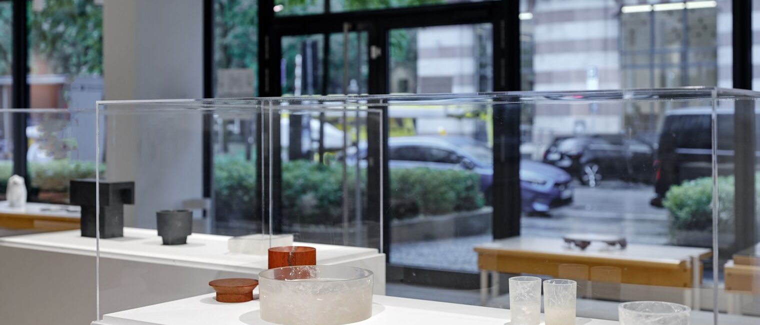 Blick durch die Galerie mit Schaukästen, die weiße Gläser, Krüge und rote und schwarze Schalen zeigen. Dahinter Schaufenster.