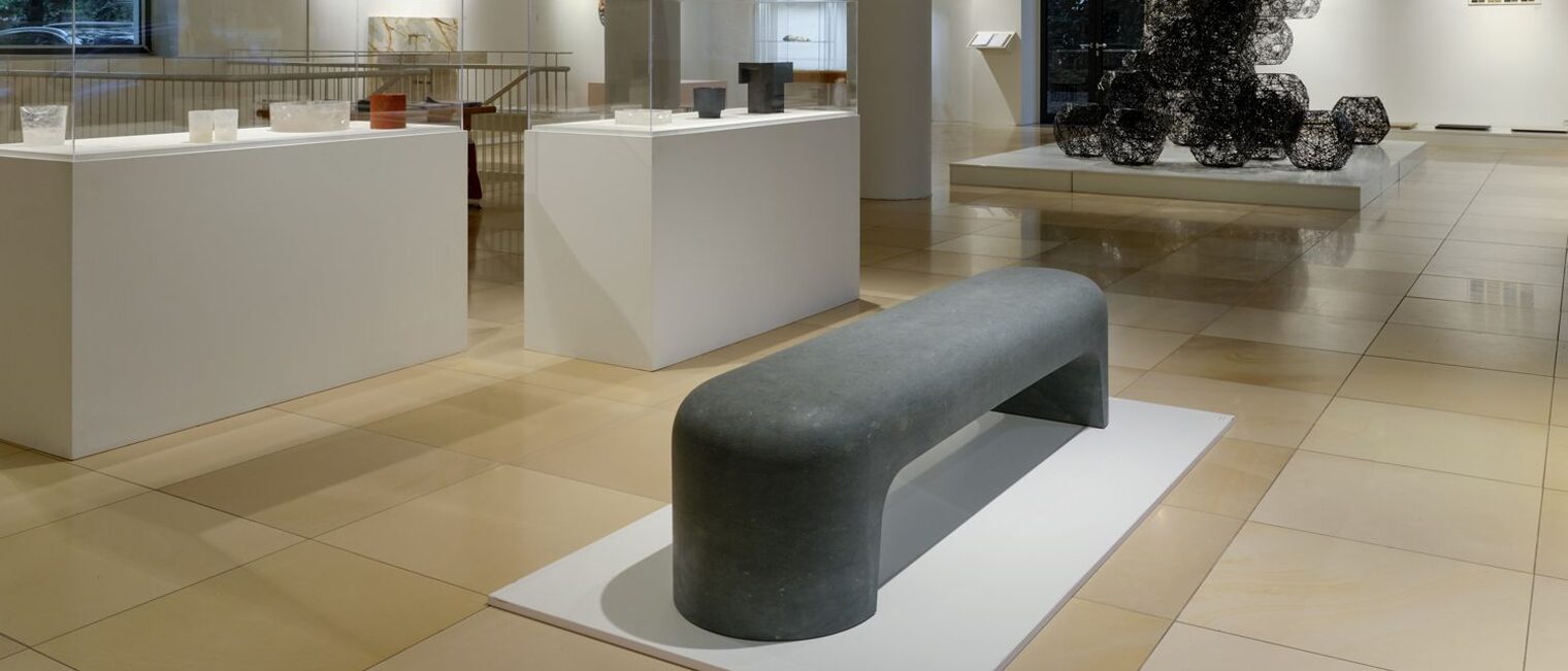 Blick durch den Ausstellungsraum. Im Vordergrund eine schwarze minimalistische Steinbank mit abgerundeten Ecken. 