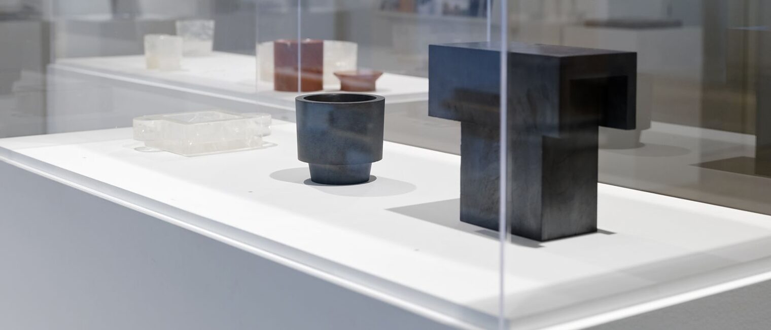 Schaukasten mit einem T-förmigen, scharfkantigen, wuchtigen Stein und ein schwarzes Trinkgefäß. Beide schwarz und glänzend.