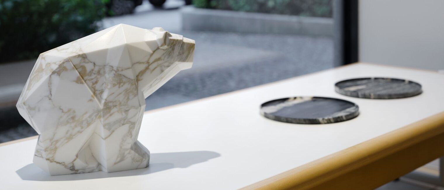 Ablage Schaufenster: ein Polarbär aus weißgrauem Marmor vor zwei schwarz-weißen Marmorplatten. Sicht auf die Büsche draußen.